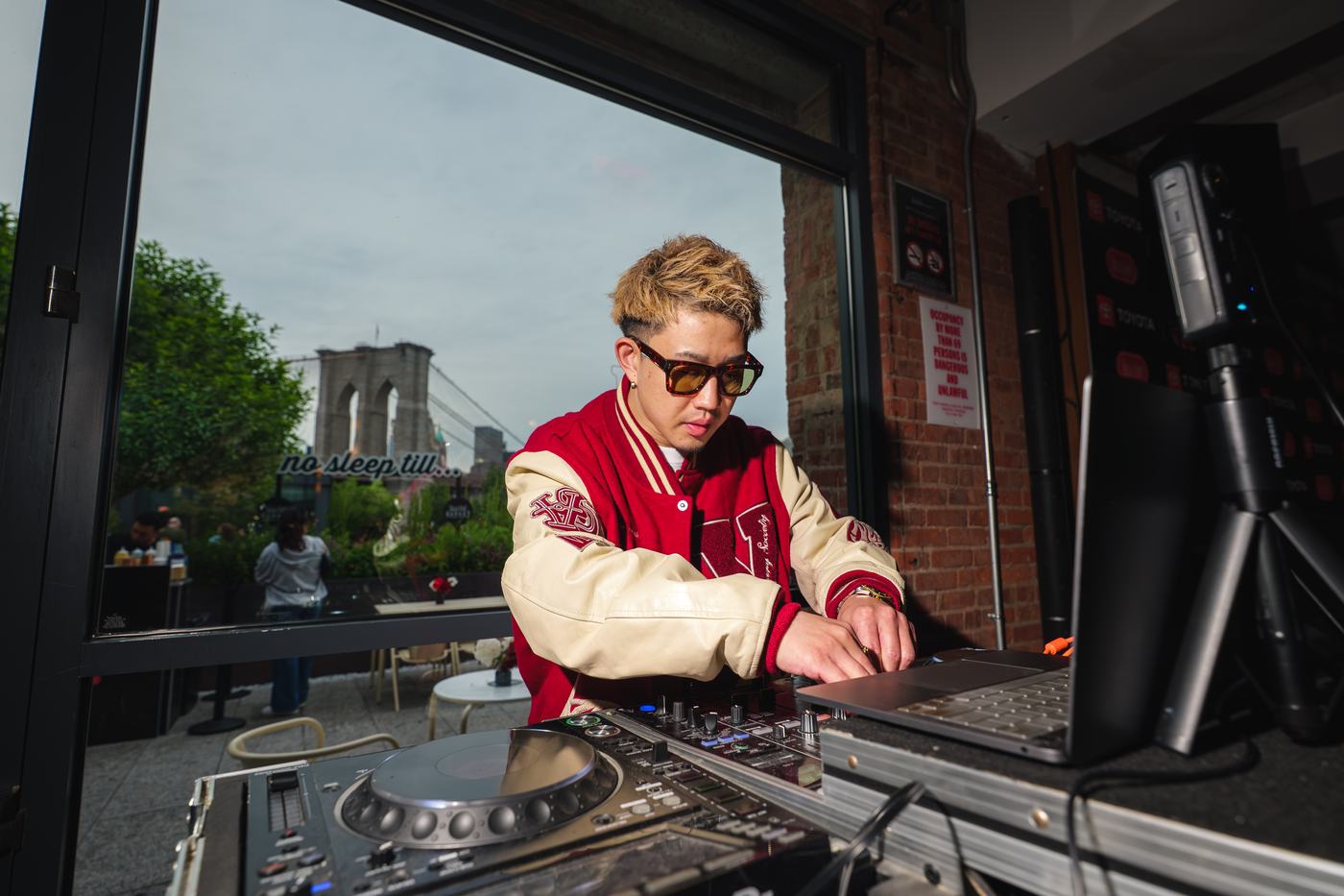 DJ performing at the activation, Brooklyn Bridge visible through the window