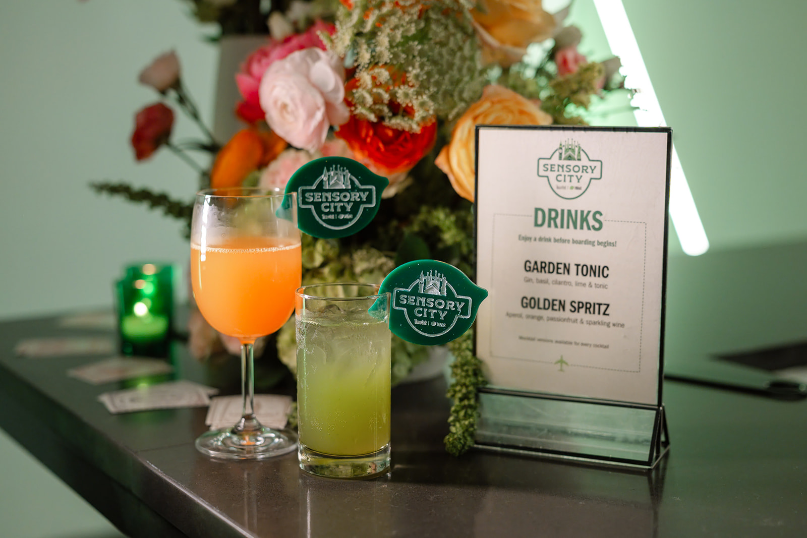 Sensory City drinks menu card beside two branded cocktails and florals